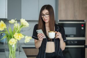Beautiful woman is using smartphone in kitchen while drinking cup of freshly brewed coffee.
