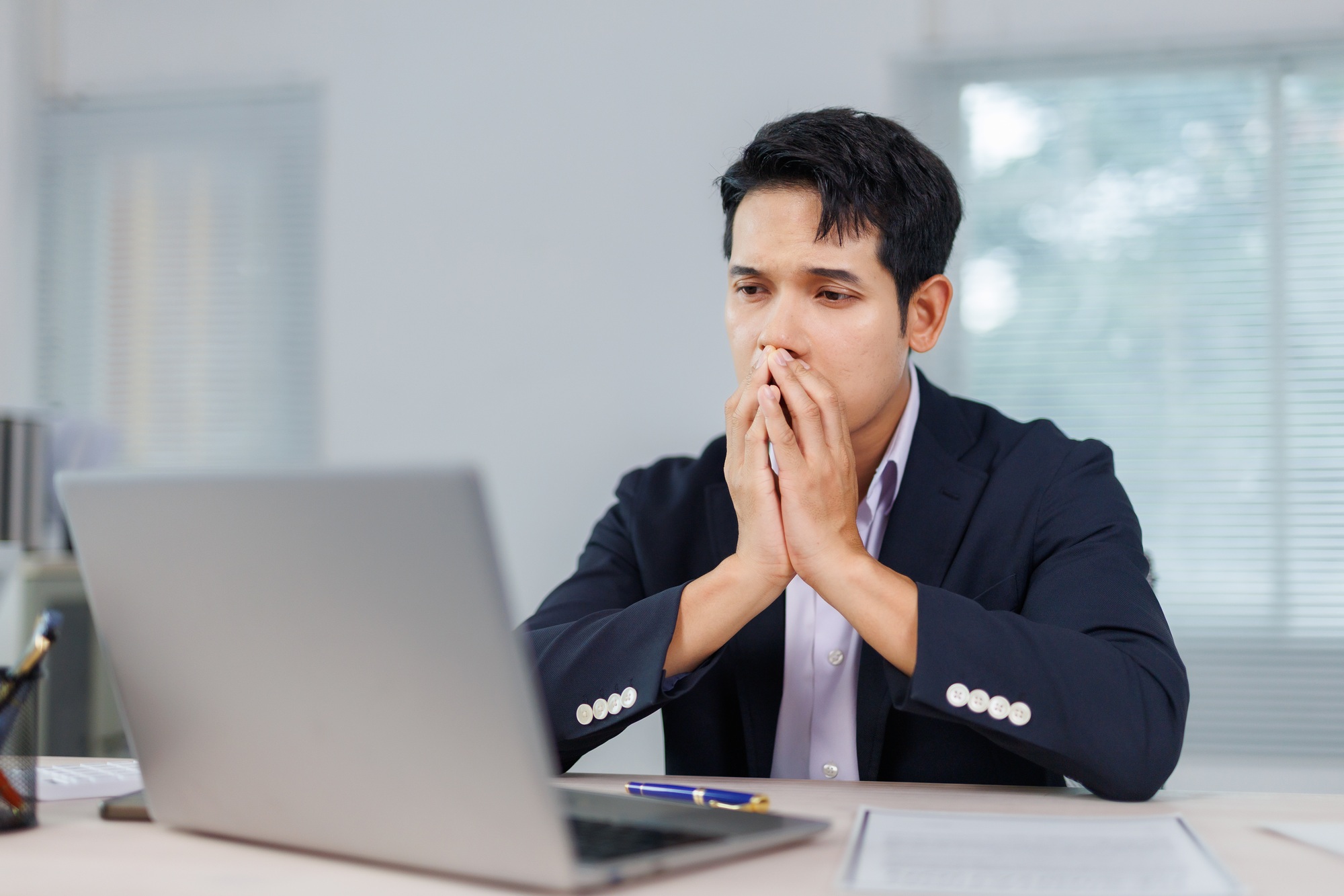 Stressed businessman covering mouth with hands while looking at laptop at office desk